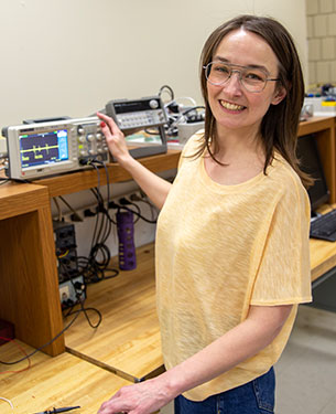 A person in a yellow top and glasses stands smiling at a workbench in a lab, holding a soldering iron. Beside them, an oscilloscope displays a waveform. The environment suggests an electronics engineering setting.