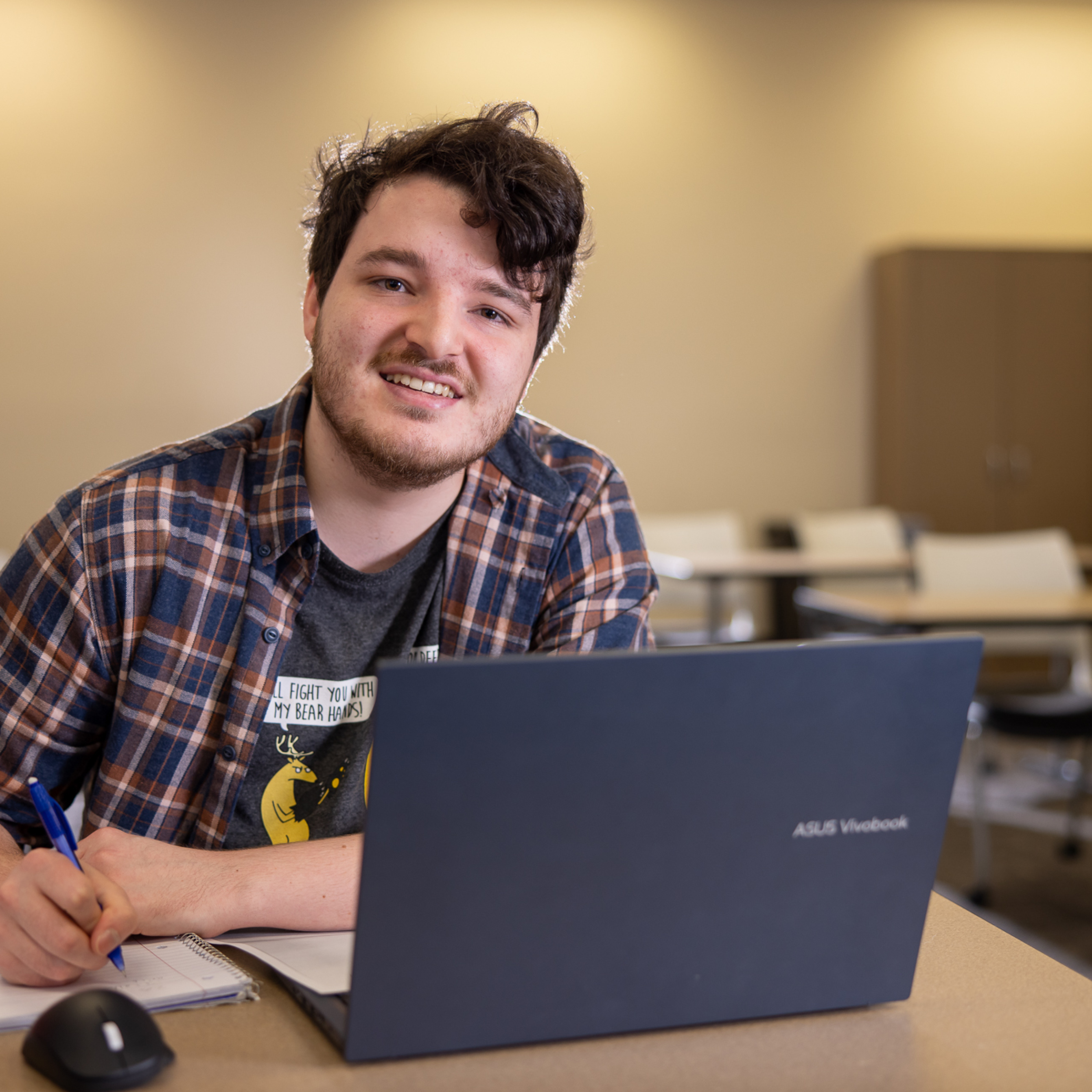 A person sits at a desk in a classroom, working on a laptop and writing in a notebook with a blue pen. 