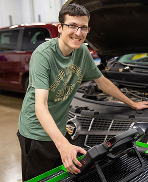 A person wearing glasses and a green t-shirt smiles while standing in front of an open car hood, working on the vehicle with a diagnostic tool in a well-lit garage.