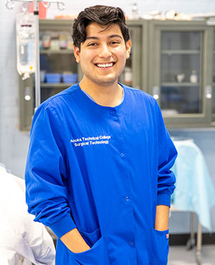 A person wearing a blue scrub top labeled "Anoka Technical College Surgical Technology" smiles broadly in a clinical laboratory setting.